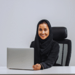 EU professional woman Amina Schulz wearing hijab, seated in ergonomic office chair with laptop, symbolizing inclusive workplace diversity in European corporate settings